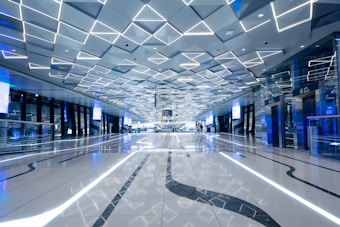 A modern, spacious interior with a geometric ceiling design featuring illuminated lines. The floor is reflective with black patterns, and there are digital screens and elevators visible. The atmosphere is futuristic and sleek with a use of blue and white lighting.