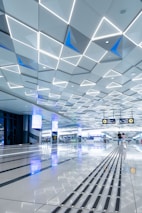 A spacious, modern interior with a geometric patterned ceiling featuring illuminated triangular shapes. The shiny floor reflects the ceiling’s lights and contains tactile guide paths. People are visible in the distance, adding a sense of scale and activity. Digital screens and signs are displayed overhead, indicating directions.