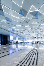A spacious, modern interior with a geometric patterned ceiling featuring illuminated triangular shapes. The shiny floor reflects the ceiling’s lights and contains tactile guide paths. People are visible in the distance, adding a sense of scale and activity. Digital screens and signs are displayed overhead, indicating directions.