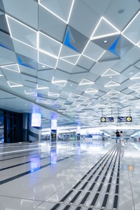 A spacious, modern interior with a geometric patterned ceiling featuring illuminated triangular shapes. The shiny floor reflects the ceiling’s lights and contains tactile guide paths. People are visible in the distance, adding a sense of scale and activity. Digital screens and signs are displayed overhead, indicating directions.