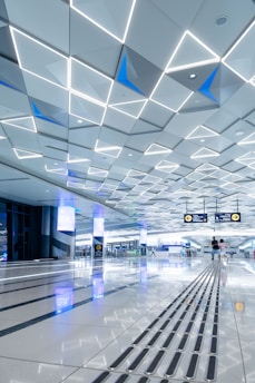 A spacious, modern interior with a geometric patterned ceiling featuring illuminated triangular shapes. The shiny floor reflects the ceiling’s lights and contains tactile guide paths. People are visible in the distance, adding a sense of scale and activity. Digital screens and signs are displayed overhead, indicating directions.
