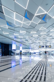 A spacious, modern interior with a geometric patterned ceiling featuring illuminated triangular shapes. The shiny floor reflects the ceiling&rsquo;s lights and contains tactile guide paths. People are visible in the distance, adding a sense of scale and activity. Digital screens and signs are displayed overhead, indicating directions.