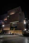 A modern, multi-level building with a geometric design features illuminated wooden panels and large recessed areas holding greenery. The lights cast strong shadows, creating an intriguing play of light and dark against the night sky. Trees in planters are placed around the structure, adding a natural element to the urban setting.