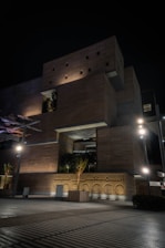 A modern, multi-level building with a geometric design features illuminated wooden panels and large recessed areas holding greenery. The lights cast strong shadows, creating an intriguing play of light and dark against the night sky. Trees in planters are placed around the structure, adding a natural element to the urban setting.