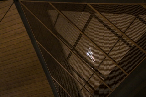 A large, abstract metal structure with geometric triangular patterns. The structure is composed of numerous parallel lines, creating a lattice-like effect. The lighting is dim, casting shadows and adding depth to the structure.