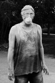 A middle-aged person with glasses and long hair is wearing a dust mask and a sleeveless, dusty shirt. They have a rugged appearance and are standing outdoors, likely engaged in some form of manual labor. The background includes trees and a road.