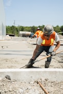 Two construction workers are smoothing out wet cement with tools on a construction site. They are wearing yellow safety vests, hard hats, and jeans. In the background, there is a large vehicle and greenery.