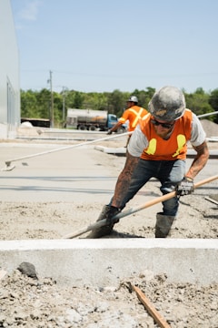 Two construction workers are smoothing out wet cement with tools on a construction site. They are wearing yellow safety vests, hard hats, and jeans. In the background, there is a large vehicle and greenery.