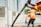 Construction workers in orange safety vests are seen pouring concrete using a long hose. The workers are focused, with one guiding the hose while another smooths the concrete surface. The setting appears to be an outdoor construction site with partially constructed walls and windows.