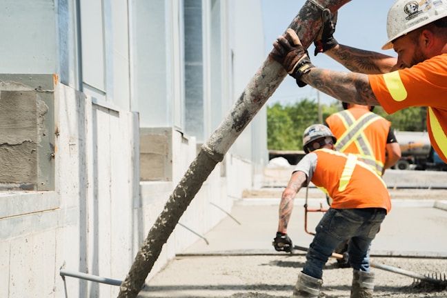Construction workers in orange safety vests are seen pouring concrete using a long hose. The workers are focused, with one guiding the hose while another smooths the concrete surface. The setting appears to be an outdoor construction site with partially constructed walls and windows.
