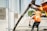 Construction workers in orange safety vests are seen pouring concrete using a long hose. The workers are focused, with one guiding the hose while another smooths the concrete surface. The setting appears to be an outdoor construction site with partially constructed walls and windows.