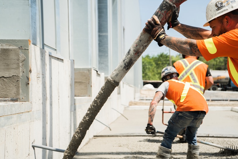 Construction workers in orange safety vests are seen pouring concrete using a long hose. The workers are focused, with one guiding the hose while another smooths the concrete surface. The setting appears to be an outdoor construction site with partially constructed walls and windows.