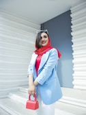 A woman with a smile stands confidently against a modern white and blue background. She is wearing a bright red hijab and a blue blazer, accessorized with a red handbag and white sunglasses resting on her head.
