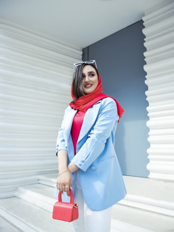 A woman with a smile stands confidently against a modern white and blue background. She is wearing a bright red hijab and a blue blazer, accessorized with a red handbag and white sunglasses resting on her head.