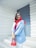 A woman with a smile stands confidently against a modern white and blue background. She is wearing a bright red hijab and a blue blazer, accessorized with a red handbag and white sunglasses resting on her head.