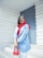 A woman with a smile stands confidently against a modern white and blue background. She is wearing a bright red hijab and a blue blazer, accessorized with a red handbag and white sunglasses resting on her head.