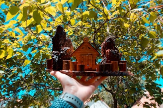 A hand wearing a blue wristband holds a small wooden model of a house with two tall tree trunks on either side. The background consists of bright green leaves with a clear turquoise sky peeking through the branches.