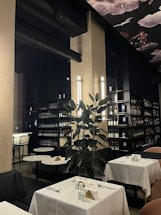 A well-lit restaurant interior featuring elegant table settings with white tablecloths and a dark, sophisticated color palette. Shelves lined with bottles of wine or liquor fill the background, hinting at a well-stocked bar area. A large leafy plant adds a natural touch, and overhead, a decorative ceiling with art depicting clouds and birds contributes to an upscale ambiance.