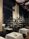 A well-lit restaurant interior featuring elegant table settings with white tablecloths and a dark, sophisticated color palette. Shelves lined with bottles of wine or liquor fill the background, hinting at a well-stocked bar area. A large leafy plant adds a natural touch, and overhead, a decorative ceiling with art depicting clouds and birds contributes to an upscale ambiance.