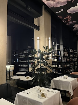 A well-lit restaurant interior featuring elegant table settings with white tablecloths and a dark, sophisticated color palette. Shelves lined with bottles of wine or liquor fill the background, hinting at a well-stocked bar area. A large leafy plant adds a natural touch, and overhead, a decorative ceiling with art depicting clouds and birds contributes to an upscale ambiance.