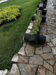 A garden scene featuring a wooden planter made from logs filled with pink and white flowers. The planter runs parallel to a stone pathway, with green grass and shrubbery on the opposite side. The stone path has a rustic, uneven appearance made of variously shaped stones.