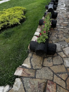 A garden scene featuring a wooden planter made from logs filled with pink and white flowers. The planter runs parallel to a stone pathway, with green grass and shrubbery on the opposite side. The stone path has a rustic, uneven appearance made of variously shaped stones.