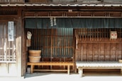 A traditional Japanese wooden storefront features horizontal slats and vertical beams. Two wooden benches are placed in front of the building, and a fabric curtain hangs above, partially obscuring the entrance. Japanese text is visible on the curtain and a vertical sign by the door. A woven basket and a large wooden bucket are among the objects present.