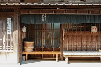 A traditional Japanese wooden storefront features horizontal slats and vertical beams. Two wooden benches are placed in front of the building, and a fabric curtain hangs above, partially obscuring the entrance. Japanese text is visible on the curtain and a vertical sign by the door. A woven basket and a large wooden bucket are among the objects present.