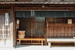 A traditional Japanese wooden storefront features horizontal slats and vertical beams. Two wooden benches are placed in front of the building, and a fabric curtain hangs above, partially obscuring the entrance. Japanese text is visible on the curtain and a vertical sign by the door. A woven basket and a large wooden bucket are among the objects present.