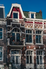 Detailed view of a historic red-brick and white-trimmed building facade featuring ornamental windows and a small balcony. The year 1894 is inscribed near the roof, with decorative elements and plants in pots on the balcony. The style reflects classic European architecture.