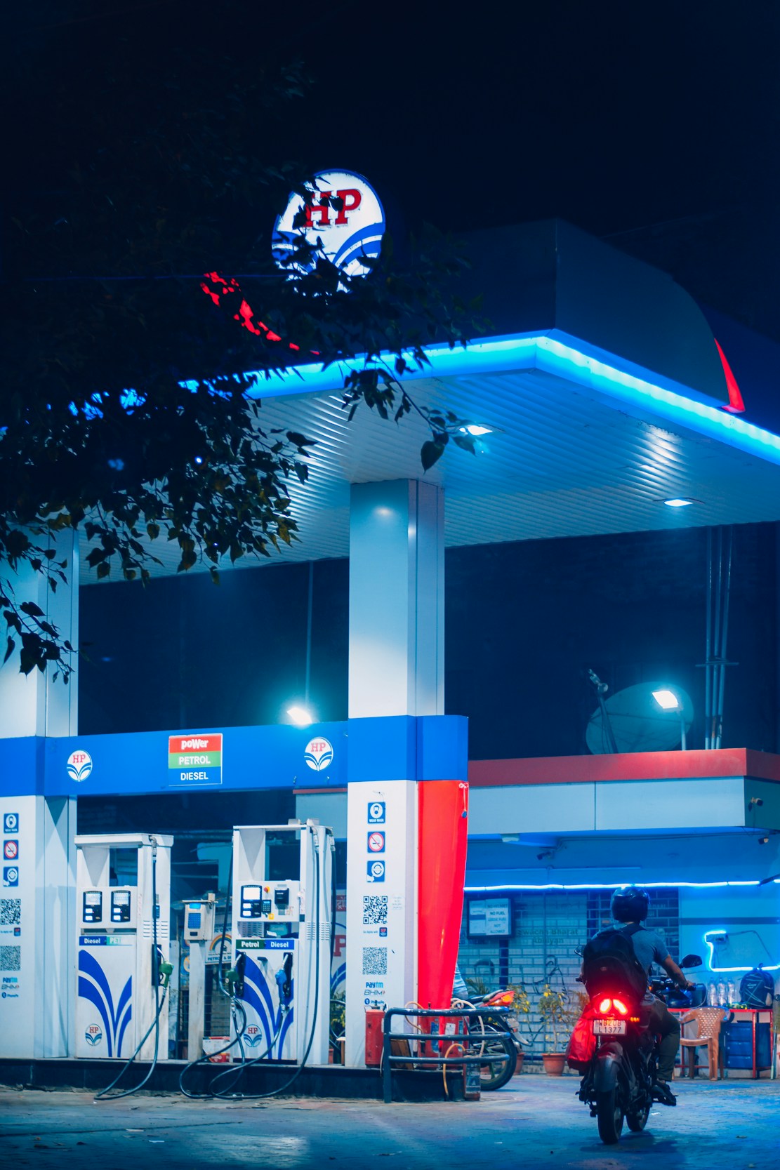 A gas station is illuminated at night, featuring bright blue and red signage and lighting. Two fuel pumps are visible, labeled for petrol and diesel. A person on a motorbike is approaching one of the pumps. The scene is partially obscured by tree leaves at the top of the image.