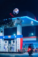 A gas station is illuminated at night, featuring bright blue and red signage and lighting. Two fuel pumps are visible, labeled for petrol and diesel. A person on a motorbike is approaching one of the pumps. The scene is partially obscured by tree leaves at the top of the image.