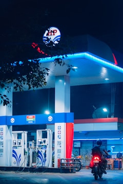 A gas station is illuminated at night, featuring bright blue and red signage and lighting. Two fuel pumps are visible, labeled for petrol and diesel. A person on a motorbike is approaching one of the pumps. The scene is partially obscured by tree leaves at the top of the image.
