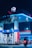 A gas station is illuminated at night, featuring bright blue and red signage and lighting. Two fuel pumps are visible, labeled for petrol and diesel. A person on a motorbike is approaching one of the pumps. The scene is partially obscured by tree leaves at the top of the image.