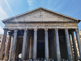 A grand ancient Roman building with tall Corinthian columns and a large triangular pediment. The inscription 'MAGRIPPALFCOSTERTIVMFECIT' is prominently displayed across the top. The structure is made of stone and has a weathered appearance, suggesting historical significance. People are gathered near the entrance, highlighting its status as a tourist attraction.
