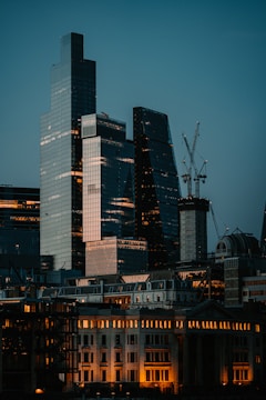 Skyscrapers with sleek glass exteriors are surrounded by construction cranes against a backdrop of a dusky sky, with older buildings at the foreground illuminated by warm lights.