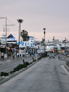 A bustling street scene features various retail and dining establishments, with people strolling along the sidewalk. A prominent building with blue-and-white accents suggests a Mediterranean architectural style. Palm trees line the street, evoking a coastal atmosphere, while an old-fashioned car drives down the road.