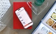 A smartphone displaying a shopping app is placed on a closed red book resting on a wooden surface. Nearby, there is an open magazine featuring watches, and a pair of eyeglasses is positioned on top of the book. A clear glass with a liquid is situated to the side.