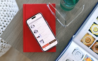 A smartphone displaying a shopping app is placed on a closed red book resting on a wooden surface. Nearby, there is an open magazine featuring watches, and a pair of eyeglasses is positioned on top of the book. A clear glass with a liquid is situated to the side.