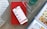 A smartphone displaying a shopping app is placed on a closed red book resting on a wooden surface. Nearby, there is an open magazine featuring watches, and a pair of eyeglasses is positioned on top of the book. A clear glass with a liquid is situated to the side.