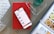 A smartphone displaying a shopping app is placed on a closed red book resting on a wooden surface. Nearby, there is an open magazine featuring watches, and a pair of eyeglasses is positioned on top of the book. A clear glass with a liquid is situated to the side.