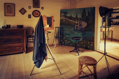 A vintage photography studio with an old wooden camera on a tripod covered by a dark cloth. The room is warmly lit and features wooden floors and furniture, including a chest of drawers and wicker stool. The backdrop depicts a painted scene of a serene lake with classical architecture. Various framed pictures are hung on the walls.