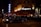 A luxurious restaurant with bright lights and a classic car parked outside. The establishment has a stylish awning and an illuminated sign. People are seen walking nearby, and the street is dimly lit, creating a sophisticated urban atmosphere.