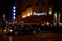 A luxurious restaurant with bright lights and a classic car parked outside. The establishment has a stylish awning and an illuminated sign. People are seen walking nearby, and the street is dimly lit, creating a sophisticated urban atmosphere.