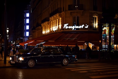A luxurious restaurant with bright lights and a classic car parked outside. The establishment has a stylish awning and an illuminated sign. People are seen walking nearby, and the street is dimly lit, creating a sophisticated urban atmosphere.