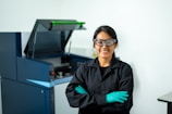 A person wearing safety goggles and a lab coat stands smiling with their arms crossed. They wear teal gloves and appear to be in a laboratory setting with equipment visible in the background, including a large machine with an open lid.
