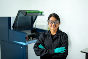 Smiling employee confidently wearing protective gloves and goggles inside a modern warehouse.