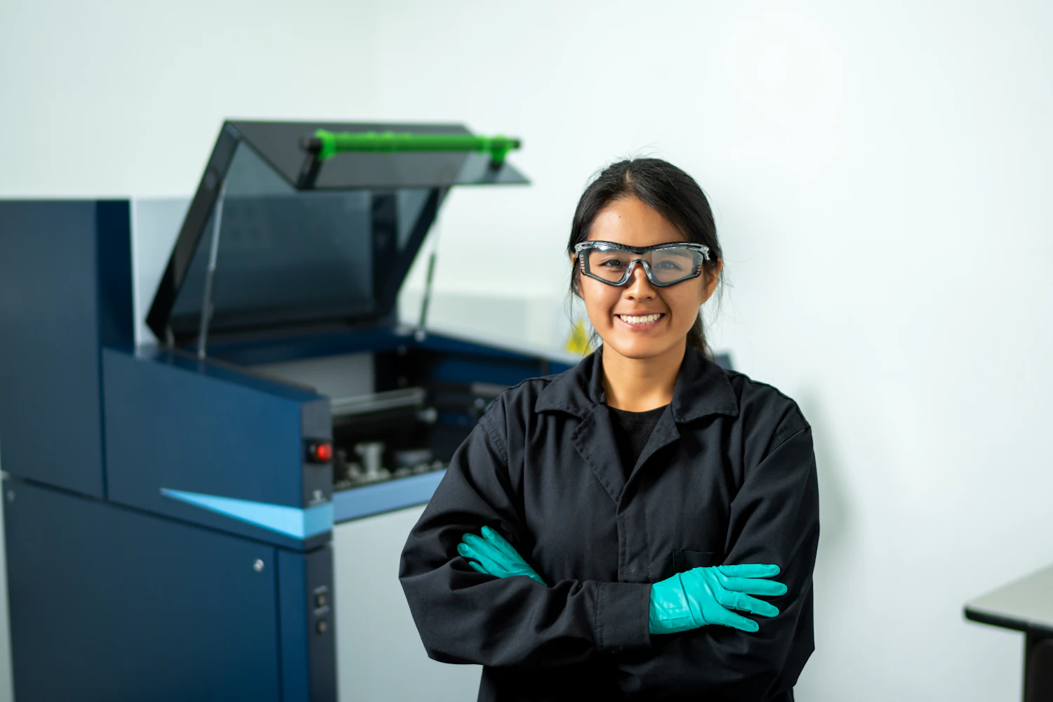 A woman in a black lab coat and safety goggles standing with arms folded beside a large press