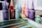 A collection of colorful poster paint bottles with pointed nozzle caps is arranged in a row. Vibrant hues include purple, red, teal, orange, pink, and yellow. In the foreground, a spattered notebook or paper displaying painted stripes in greens, blues, and browns is visible.