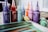 A collection of colorful poster paint bottles with pointed nozzle caps is arranged in a row. Vibrant hues include purple, red, teal, orange, pink, and yellow. In the foreground, a spattered notebook or paper displaying painted stripes in greens, blues, and browns is visible.
